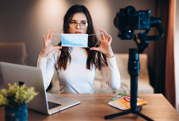 La joven bloguera con gafas está filmando su vlog y mostrando a su audiencia que esté segura y use una máscara médica. Concepto de lugar de trabajo seguro de coronovirus. Ponerse una mascarilla médica en la cara.