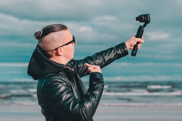 Joven bloguera con gafas de sol haciendo Selfie o transmisión de video en la playa con cámara de acción con estabilizador de cámara Gimbal.