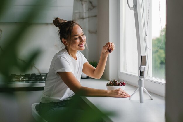 Una joven bloguera nutricionista realiza una consulta o una lección abierta mediante un enlace de vídeo que habla de los beneficios de las bayas frescas sentadas en la cocina con un plato de cerezas