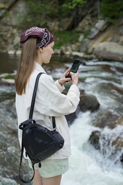 Una joven bloguera que usa una chaqueta y pantalones de mezclilla blanca está filmando un río rápido y un acantilado en su teléfono El concepto de unidad con la naturaleza viajar en solitario