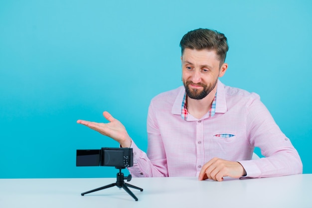 El joven bloguero está posando para su mini cámara apuntando a la izquierda con la mano en el fondo azul