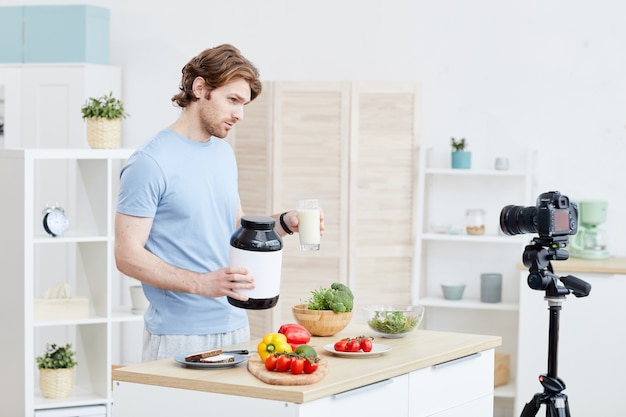 Joven bloguero hablando sobre comida deportiva y alimentación saludable en cámara para su blog mientras está de pie en la cocina