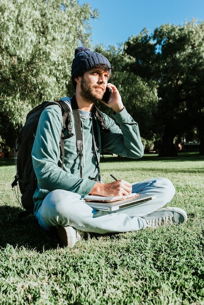 Un joven bloguero que escribe en su bloc de notas disfruta de la vida nómada digital. Habla por teléfono con sus clientes mientras toma notas. Concepto: felicidad y libertad