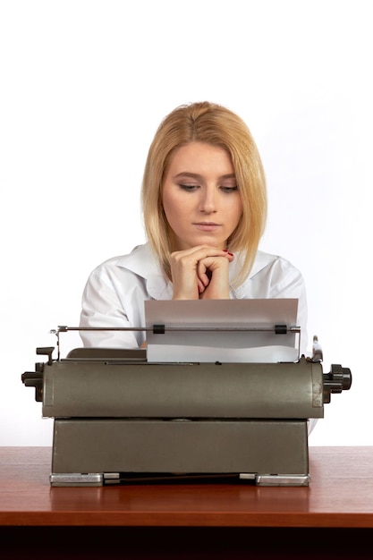 Una joven con una blusa blanca se sienta en un escritorio en la oficina y escribe texto en una máquina de escribir retro.