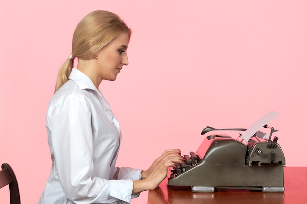 Una joven con una blusa blanca se sienta en un escritorio en la oficina y escribe texto en una máquina de escribir retro.