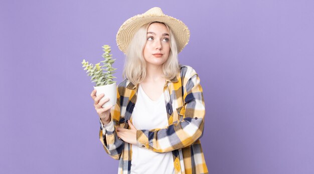 Joven bonita albina encogiéndose de hombros, sintiéndose confundida e insegura y sosteniendo un cactus de planta de interior