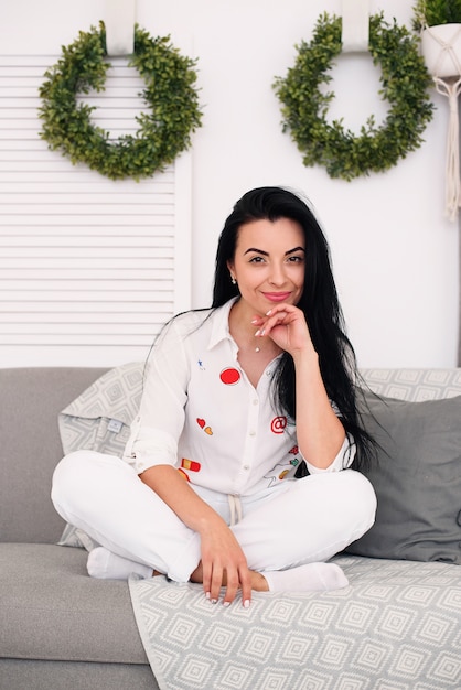 Joven bonita chica morena descansar en una acogedora habitación luminosa. Mujer sentada en el sofá gris en interior escandinavo.