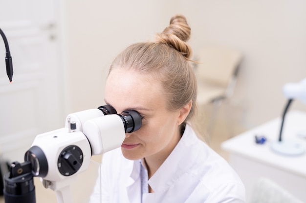 Joven bonita a doctor de la vista mirando a través de equipos optométricos mientras examina la vista del paciente en clínicas