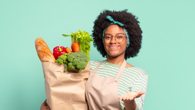 Joven bonita mujer afro que parece feliz, asombrada y sorprendida, sonriendo y dándose cuenta de buenas noticias increíbles e increíbles y sosteniendo una bolsa de verduras