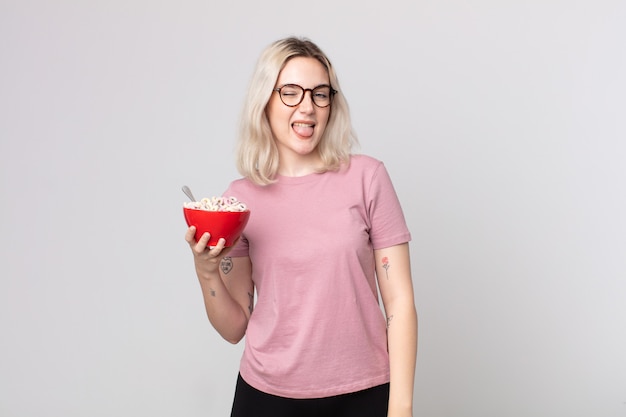 Joven bonita a mujer albina con actitud alegre y rebelde, bromeando y sacando la lengua con un tazón de desayuno