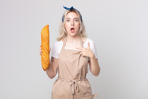 Joven bonita a mujer albina mirando conmocionado y sorprendido con la boca abierta, apuntando a sí mismo con una baguette de pan