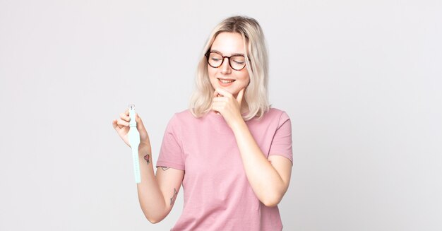 Joven bonita a mujer albina sonriendo con una expresión feliz y segura con la mano en la barbilla sosteniendo un reloj reloj