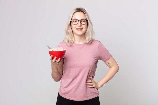 Joven bonita a mujer albina sonriendo felizmente con una mano en la cadera y confiada con un tazón de desayuno