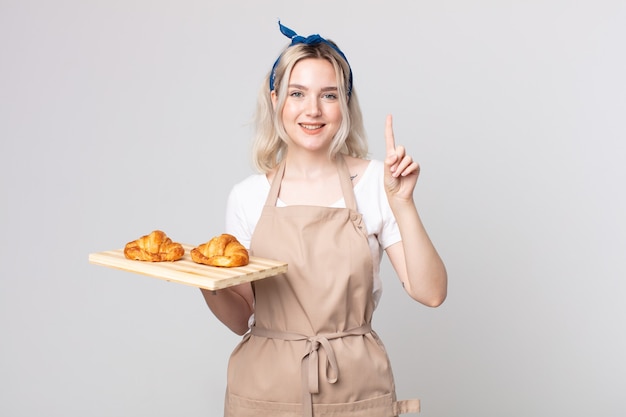 Joven bonita a mujer albina sonriendo y mirando amigable, mostrando el número uno con una bandeja de croissants