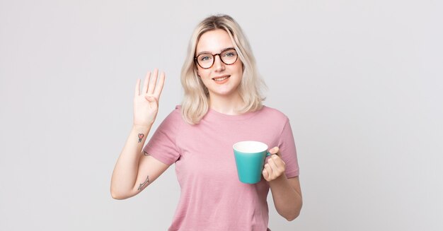 Joven bonita a mujer albina sonriendo y mirando amigable, mostrando el número cuatro con una taza de café