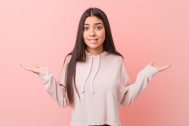 Joven y bonita mujer árabe vestida con un look casual deportivo confundida y dudosa encogiéndose de hombros para sostener un espacio de copia.