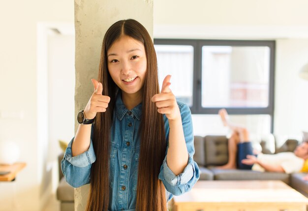 Joven bonita a mujer asiática sonriendo con una actitud positiva, exitosa y feliz apuntando al frente, haciendo un signo de pistola con las manos