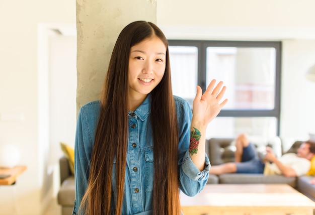 Joven bonita a mujer asiática sonriendo feliz y alegremente, saludando con la mano, dándote la bienvenida y saludándote, o diciéndote adiós