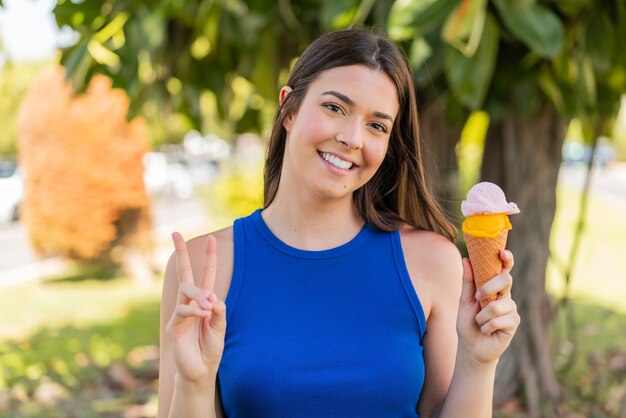 Joven y bonita mujer brasileña con un helado de cucurucho al aire libre sonriendo y mostrando el signo de la victoria