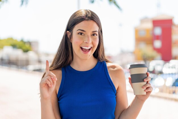 Joven y bonita mujer brasileña sosteniendo un café para llevar al aire libre señalando una gran idea