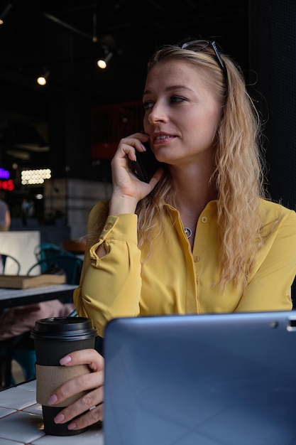 Joven bonita a mujer caucásica en la cafetería con ordenador portátil llamando por teléfono inteligente