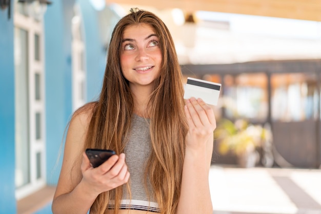 Joven y bonita mujer caucásica comprando con el móvil con una tarjeta de crédito mientras piensa