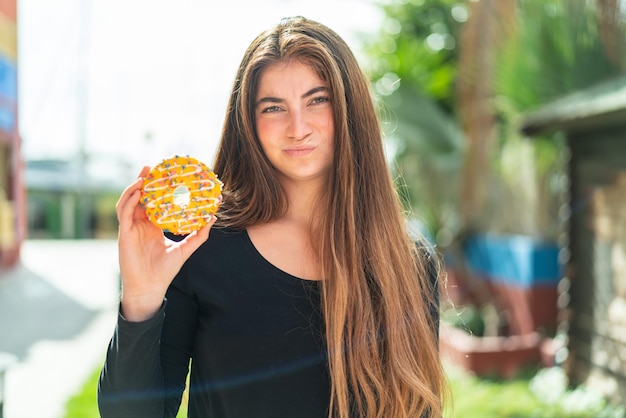 Joven y bonita mujer caucásica sosteniendo una rosquilla al aire libre con expresión triste