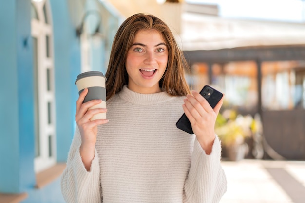 Joven y bonita mujer caucásica usando un teléfono móvil y sosteniendo un café con expresión feliz