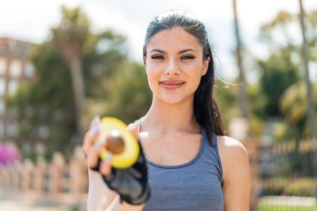 Joven bonita mujer deportiva sosteniendo un aguacate al aire libre con expresión feliz
