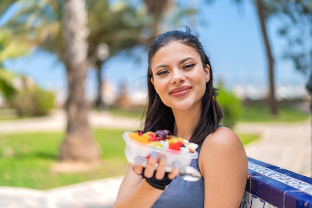 Foto joven bonita mujer deportiva sosteniendo un tazón de fruta al aire libre con expresión feliz