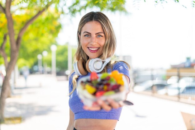 Foto joven bonita mujer deportiva sosteniendo un tazón de fruta al aire libre con expresión feliz