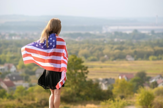 Joven y bonita mujer estadounidense con cabello largo agitando la bandera de los Estados Unidos en el viento en sus hombros de pie al aire libre disfrutando del cálido día de verano