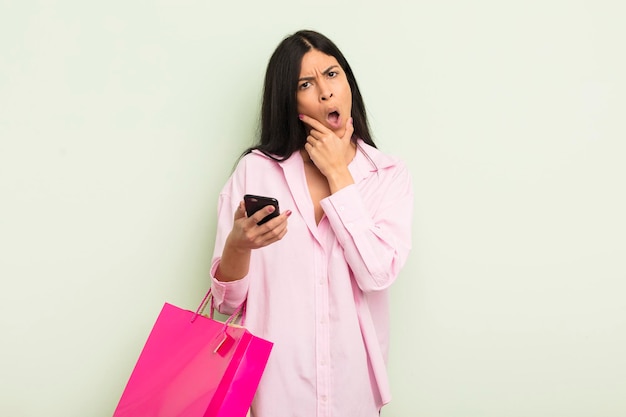 Joven bonita mujer hispana con la boca y los ojos bien abiertos y con la mano en la barbilla concepto de bolsas de compras