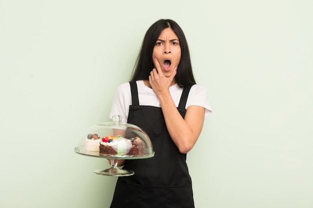 Joven bonita mujer hispana con la boca y los ojos bien abiertos y la mano en el mentón chef con concepto de pasteles