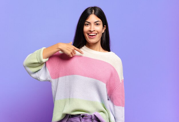 Joven bonita a mujer hispana que parece feliz, orgullosa y sorprendida, apuntando alegremente a sí misma, sintiéndose segura y elevada