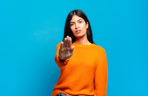 Joven bonita a mujer hispana que parece seria, severa, disgustada y enojada mostrando la palma abierta haciendo gesto de parada