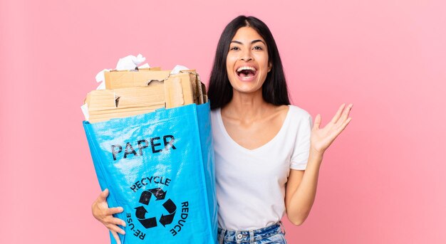 Joven bonita a mujer hispana sintiéndose feliz y asombrada por algo increíble y sosteniendo una bolsa de papel para reciclar