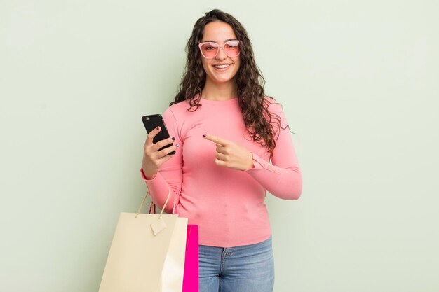 Joven bonita mujer hispana sonriendo alegremente sintiéndose feliz y señalando el concepto de bolsas de compras laterales