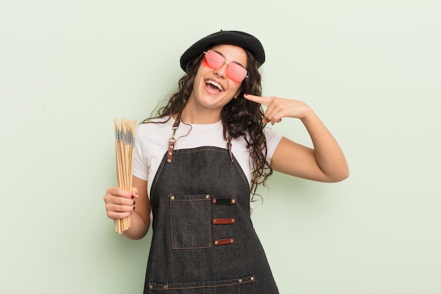 Joven bonita mujer hispana sonriendo con confianza apuntando a su propio concepto de artista de sonrisa amplia