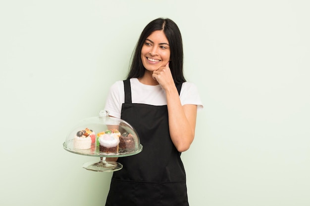 Joven bonita mujer hispana sonriendo con una feliz expresión de confianza con la mano en la barbilla chef con concepto de pasteles