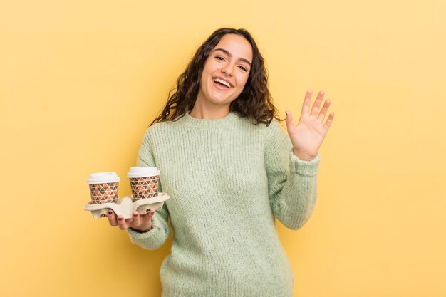 Joven bonita mujer hispana sonriendo felizmente agitando la mano dándole la bienvenida y saludando el concepto de café para llevar