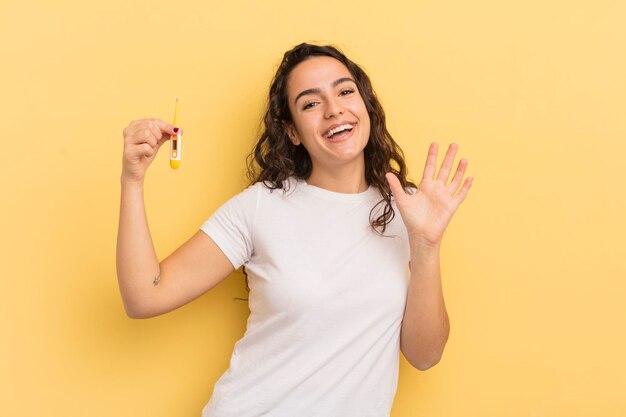 Joven bonita mujer hispana sonriendo felizmente agitando la mano dándole la bienvenida y saludando el concepto de termómetro