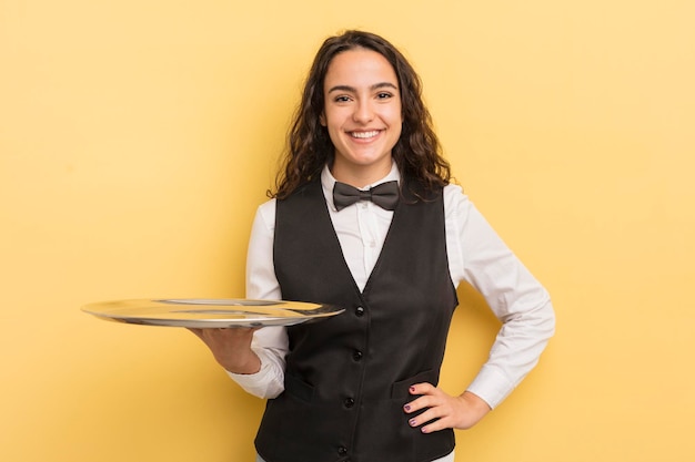 Joven bonita mujer hispana sonriendo felizmente con una mano en la cadera y confiado concepto de camarero y bandeja