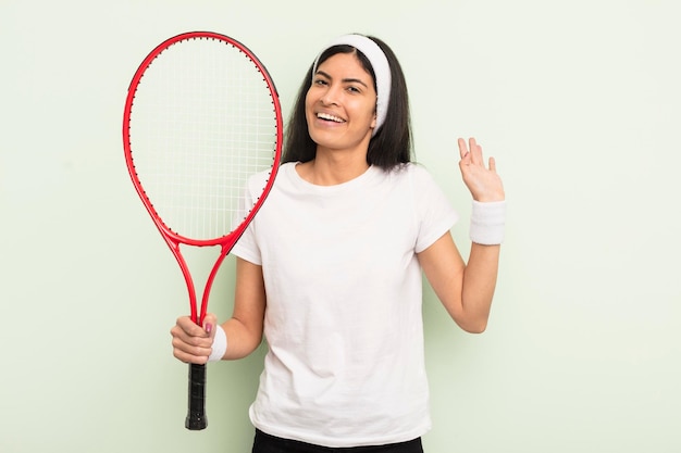 Joven bonita mujer hispana sonriendo felizmente saludando con la mano dándole la bienvenida y saludando el concepto de tenis