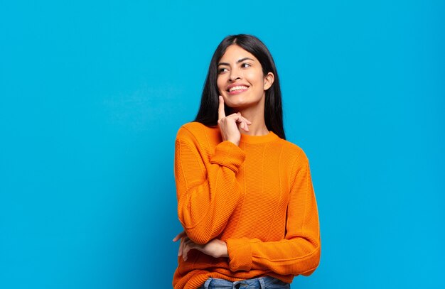 Joven bonita a mujer hispana sonriendo felizmente y soñando despierto o dudando, mirando hacia el lado