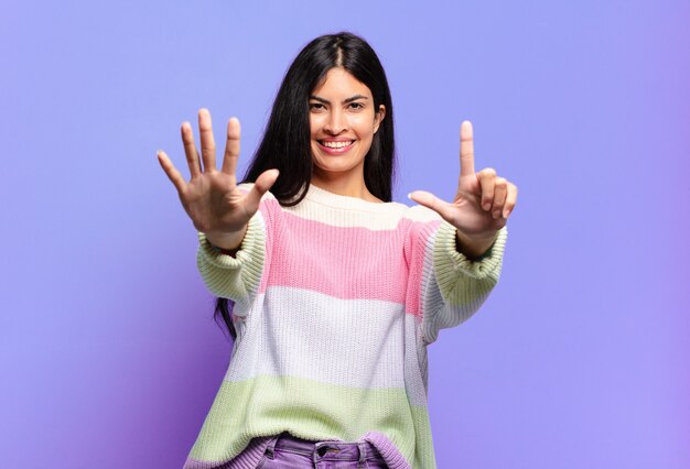 Joven bonita a mujer hispana sonriendo y mirando amistosamente, mostrando el número siete o séptimo con la mano hacia adelante, contando hacia atrás