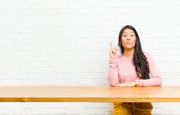 Joven y bonita mujer latina que se siente como un genio sosteniendo el dedo con orgullo en el aire después de darse cuenta de una gran idea, diciendo eureka sentada frente a una mesa