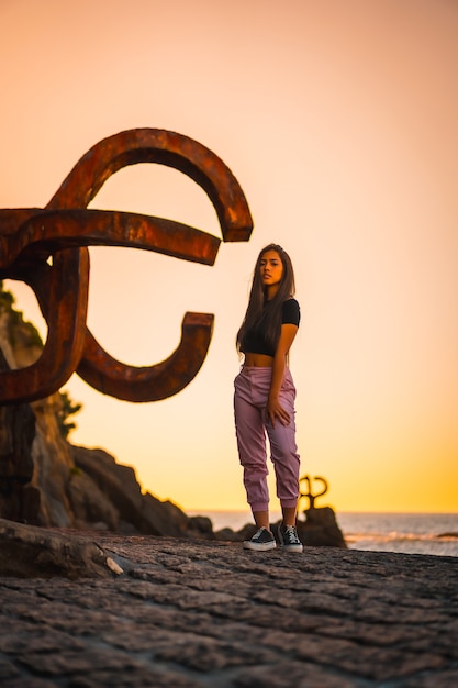 Una joven y bonita mujer morena de pelo largo y liso con una camiseta negra corta junto a una famosa escultura de San Sebatian llamada Peine del Viento