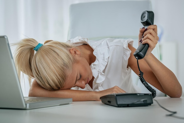 Una joven y bonita mujer de negocios, agotada por el trabajo, apoyada en el escritorio de la oficina con un teléfono en la mano y durmiendo después de las malas noticias que escuchó.