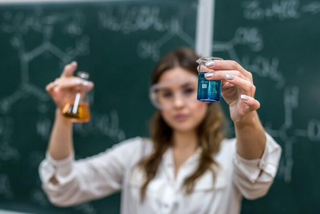 Foto joven bonita profesora sostiene el matraz con el líquido de color cerca de la pizarra en una lección de química en clase.